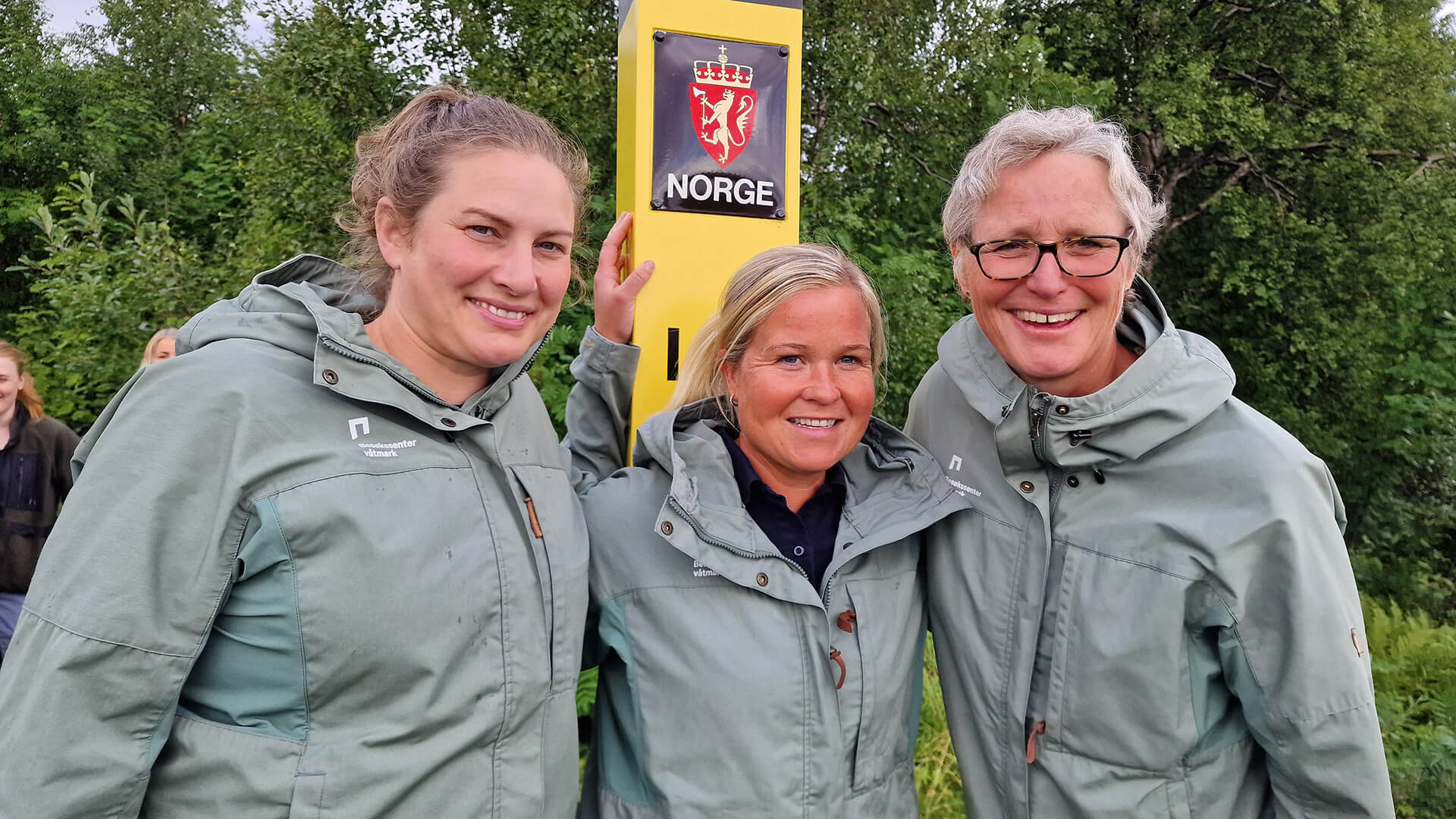 Tre personer ved gul grensestolpe i Pasvik.